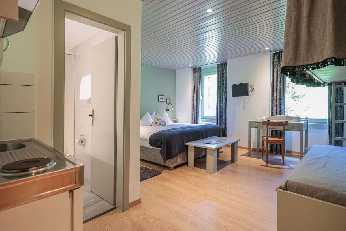 Ein Schlafzimmer mit einem Bett, Nachttisch und Schreibtisch mit Stuhl. Ein Fenster mit Vorhängen bietet Blick ins Grüne.