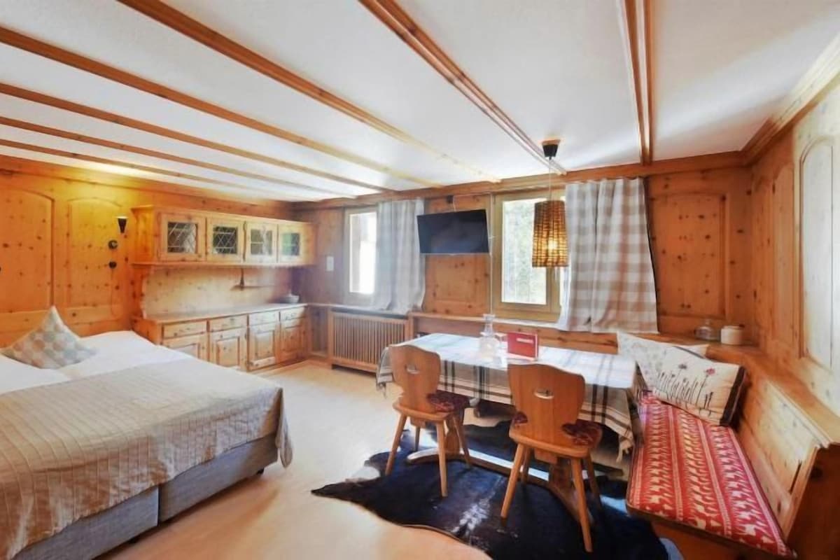 A wooden cabin interior with a bed, a dining table with chairs, and a bench.