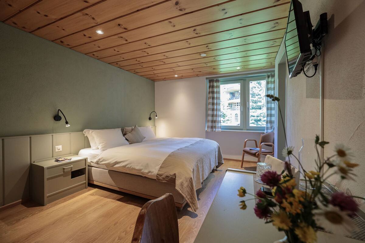 Ein Schlafzimmer mit Holzbalkendecke, einem Bett, einem Nachttisch, einem Schreibtisch, einem Stuhl und einem Fenster mit Blick auf Gebäude.