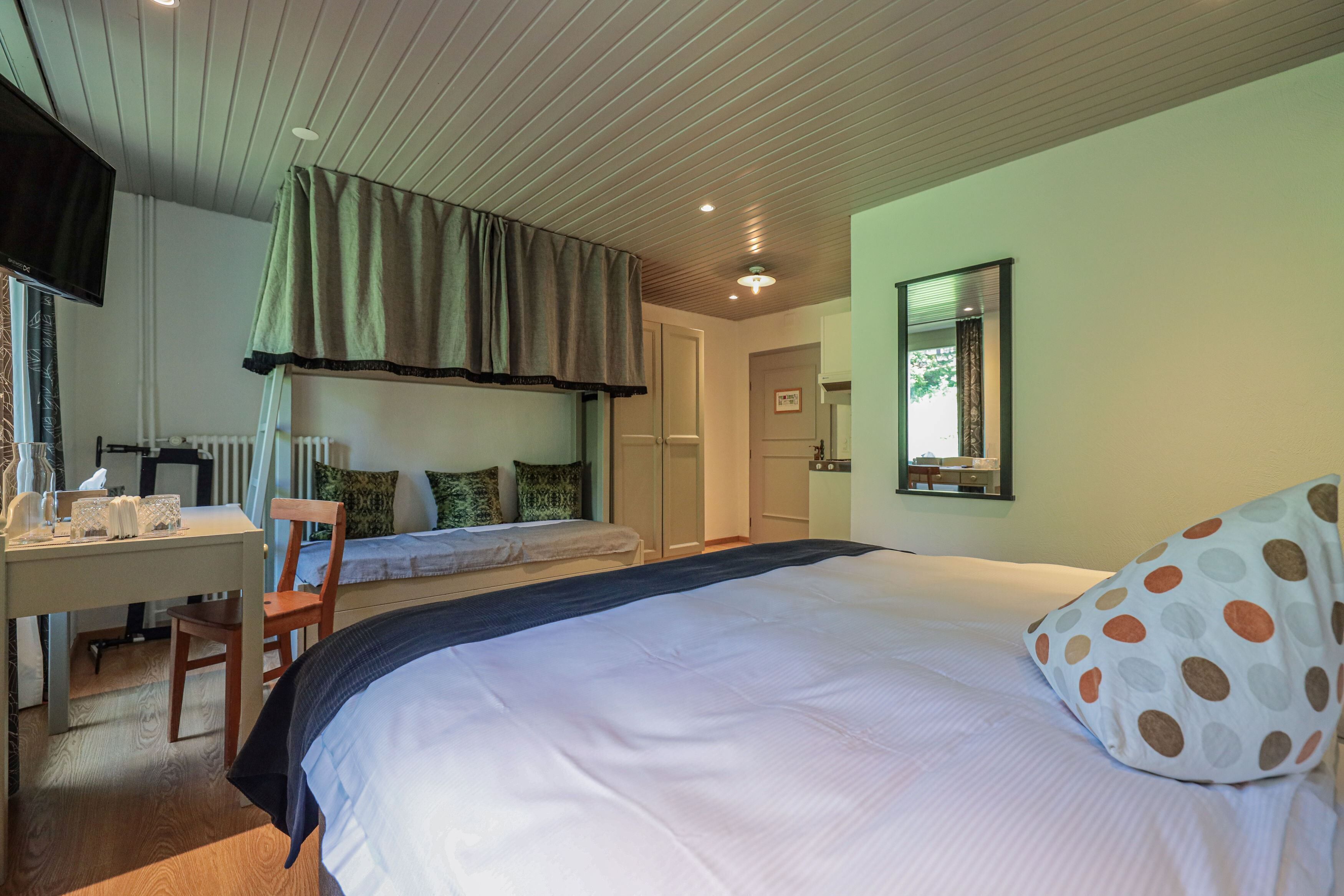 A bedroom with a large bed, a desk, and a chair. There is a window with curtains, a door, and a mirror on the wall.