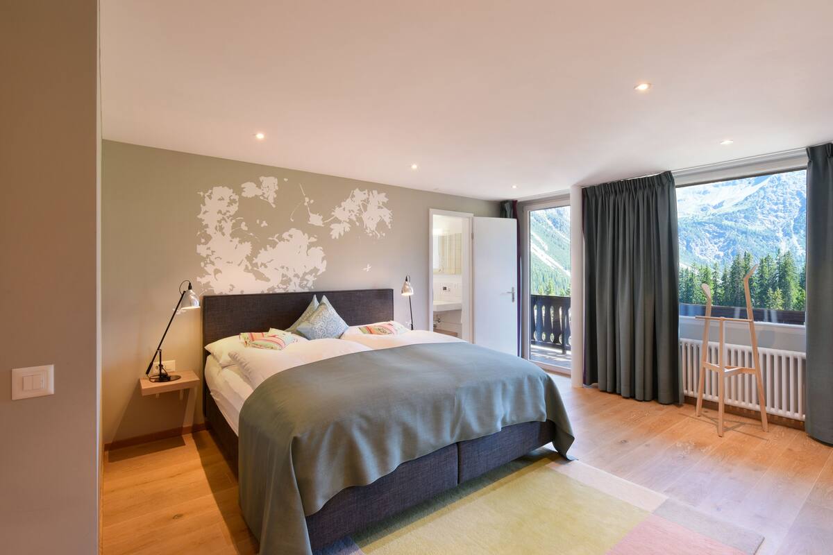 Ein Schlafzimmer mit Bett, Nachttisch, Lampe und Blick auf Berge vom Balkon aus.