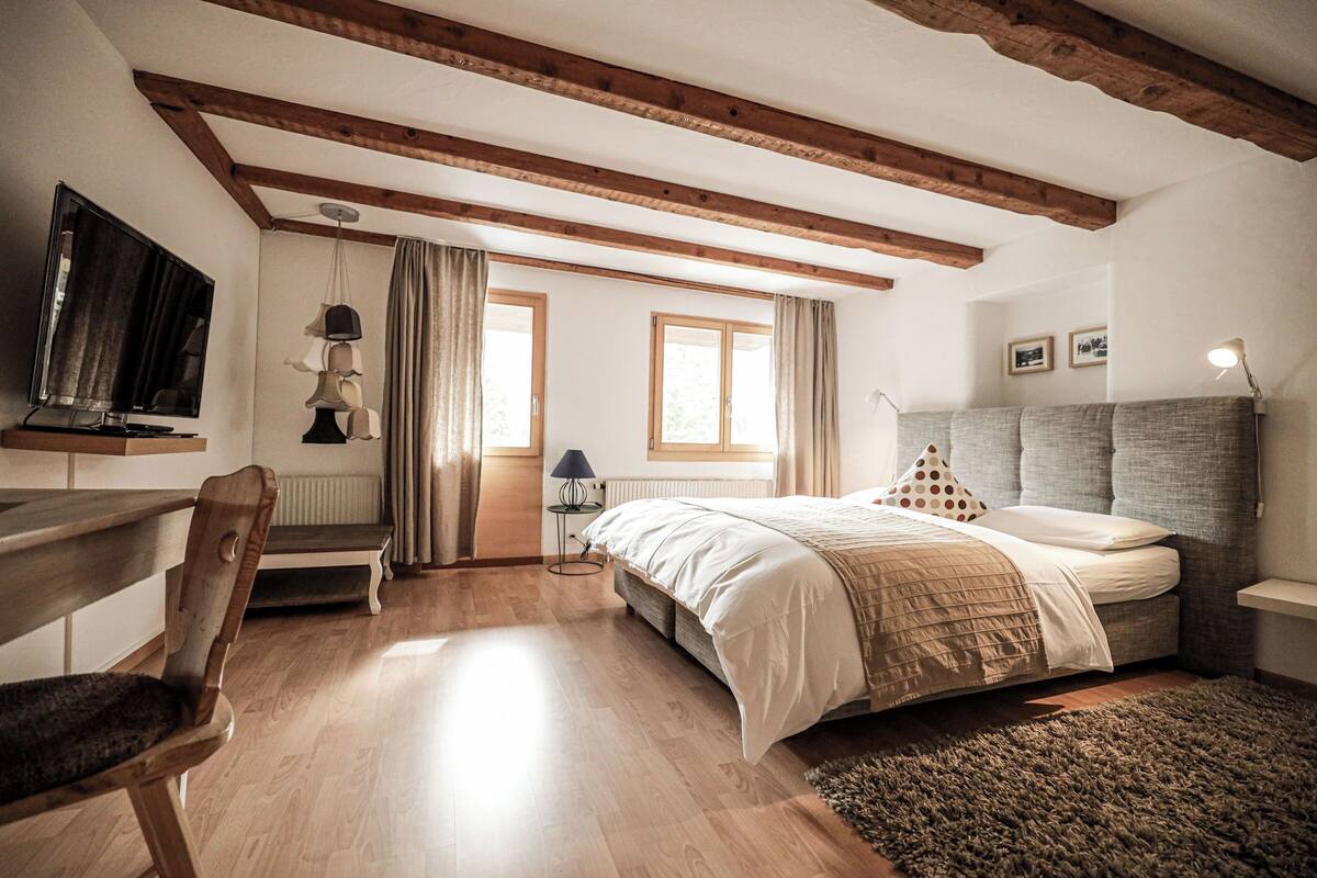 Ein Schlafzimmer mit Holzbalkendecke, einem Bett mit grauem Kopfteil, einem Schreibtisch mit Stuhl, einem Fernseher und einem Fenster mit Vorhängen.