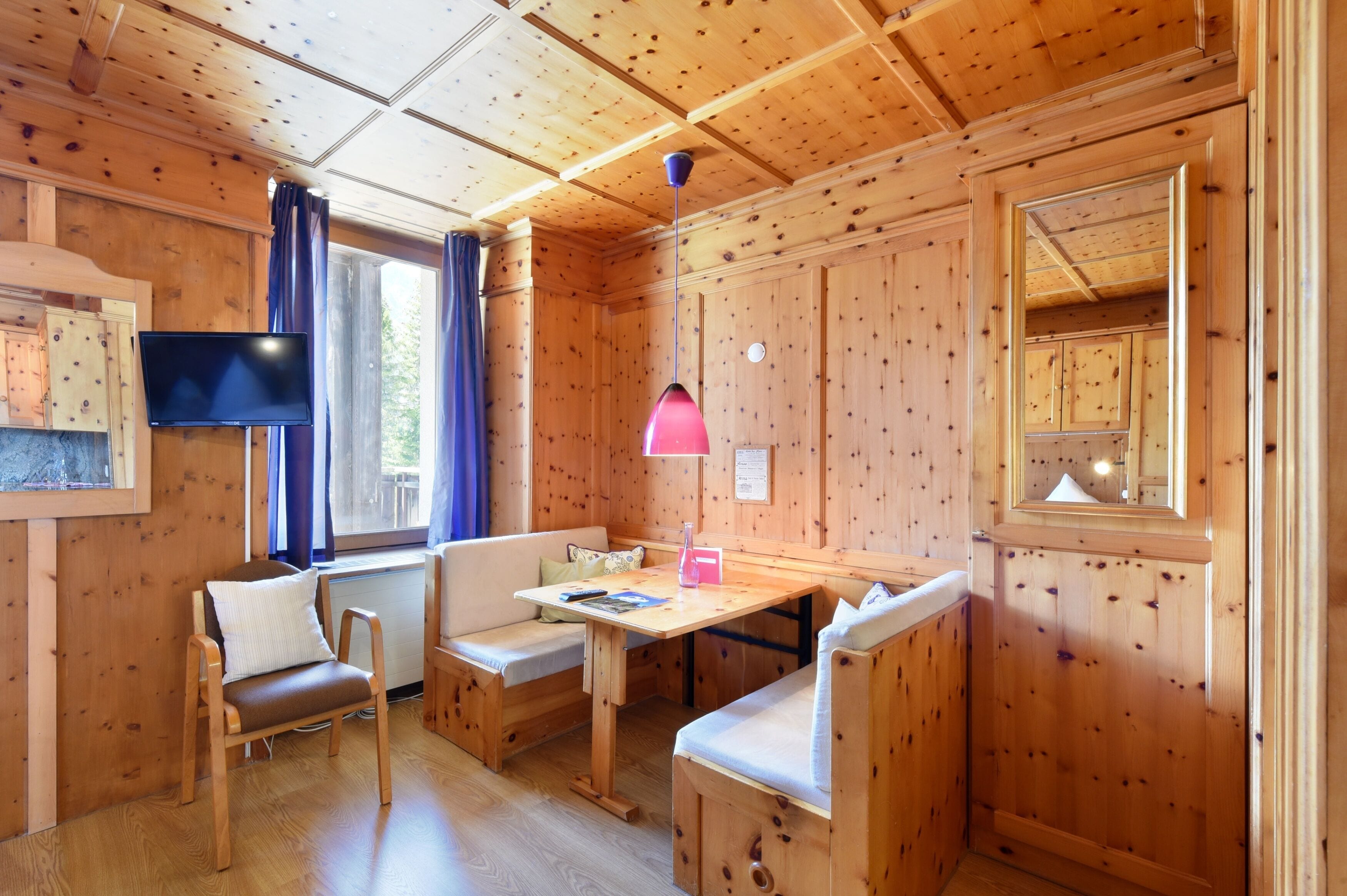 A cozy cabin interior with wooden walls, a wooden table, two cushioned benches, a flat-screen TV, and a window with curtains.