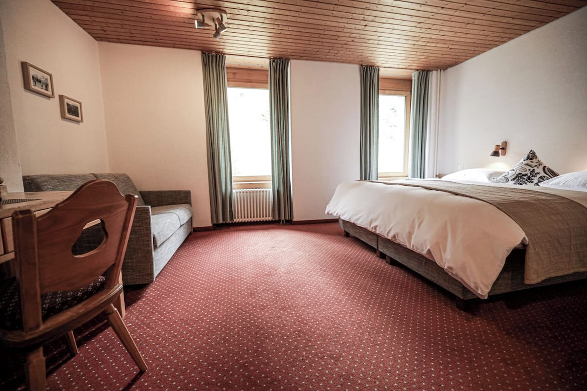 Ein Schlafzimmer mit Bett, Schreibtisch und Stuhl. Zwei Fenster mit Vorhängen.