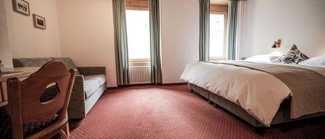A bedroom with a bed, a desk, and a chair. There are two windows with curtains.