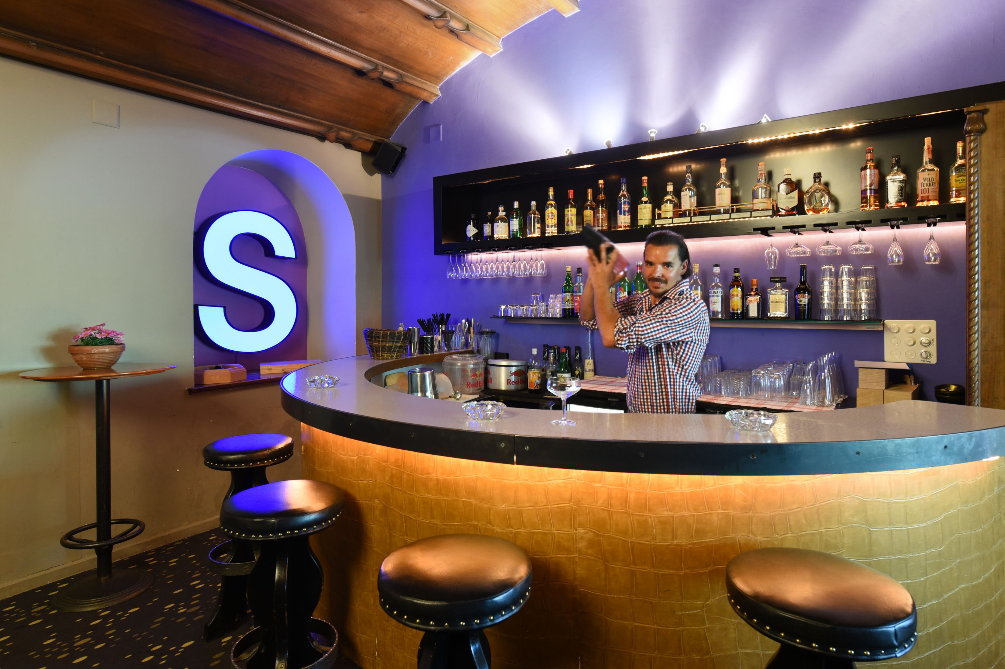 A bar area with a bartender, a large illuminated 'S' sign, and a variety of bottles and glasses on shelves.
