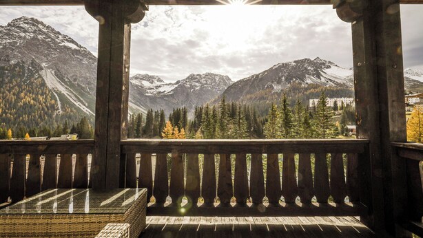 Un balcón de madera con una mesa y banco de vidrio, con vistas a un paisaje montañoso con picos nevados y una zona boscosa.