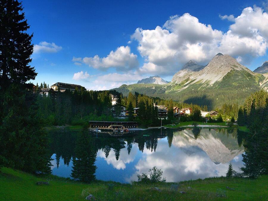 A lakeside view with a mountain in the background, a dock with boats, and buildings surrounded by trees.