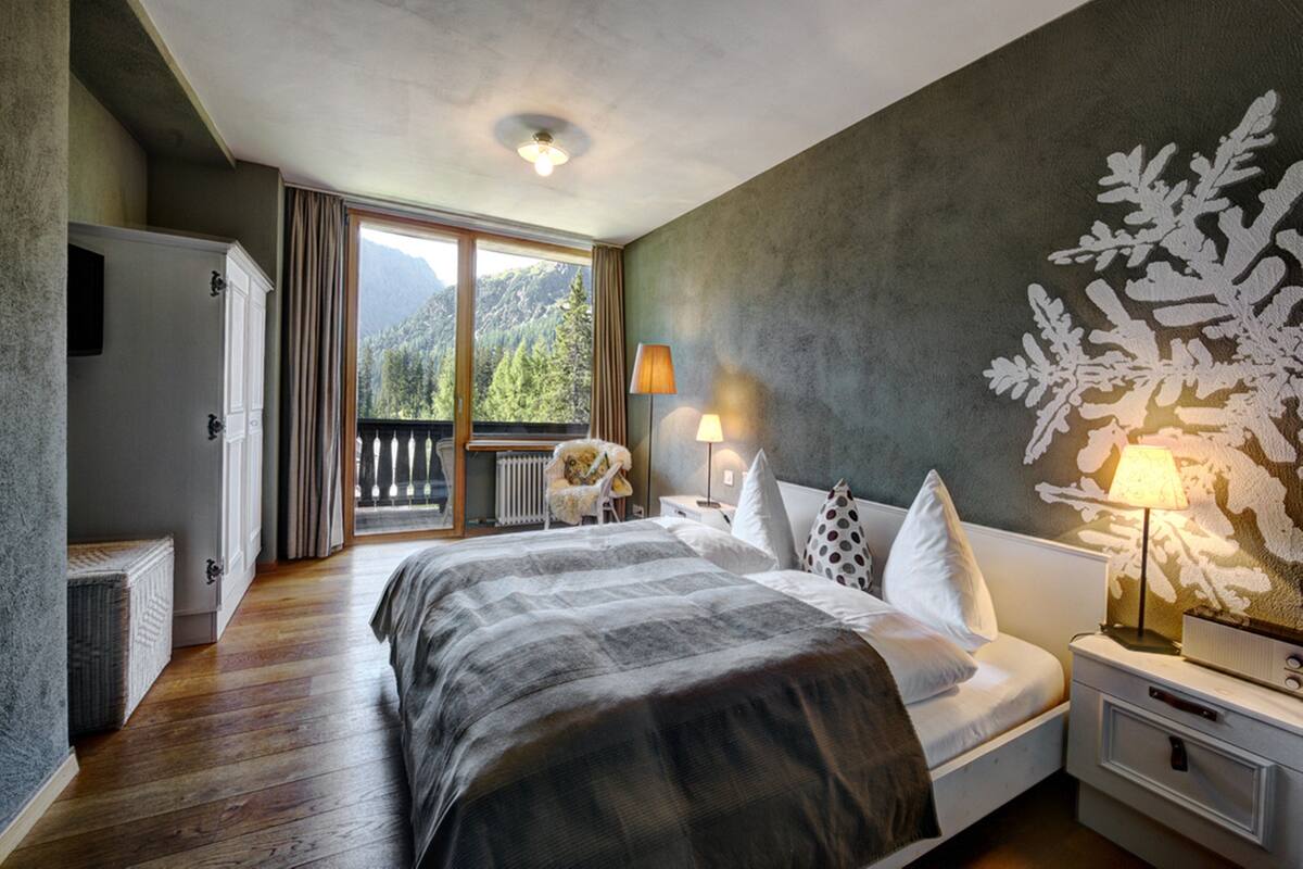 Ein Schlafzimmer mit einem großen Bett, einem Nachttisch mit Lampen, Blick auf Berge durch eine Schiebetür und einem Heizkörper unter der Tür.