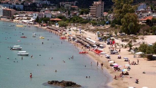 Una playa con gente nadando y tomando el sol, barcos en el agua y edificios al fondo.