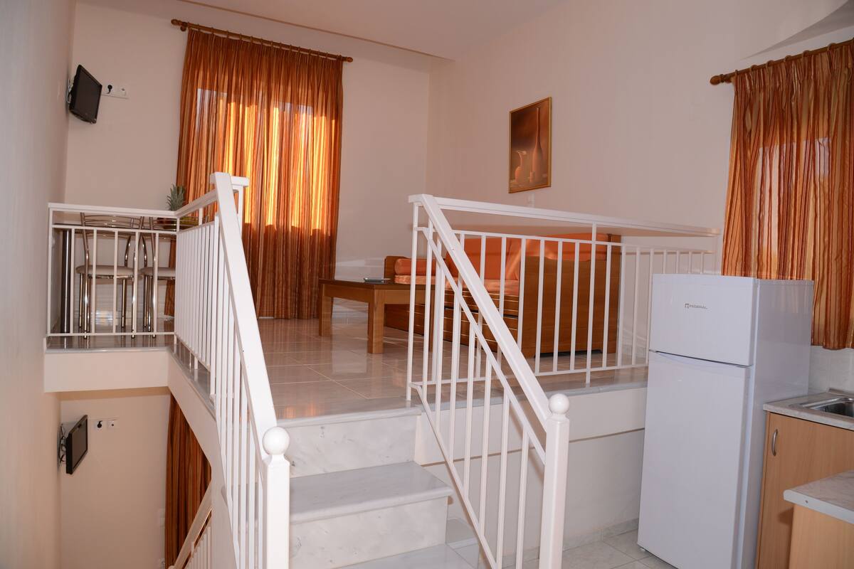 A kitchen with a refrigerator, a dining table, and chairs. There is a staircase with a railing leading to an upper level.