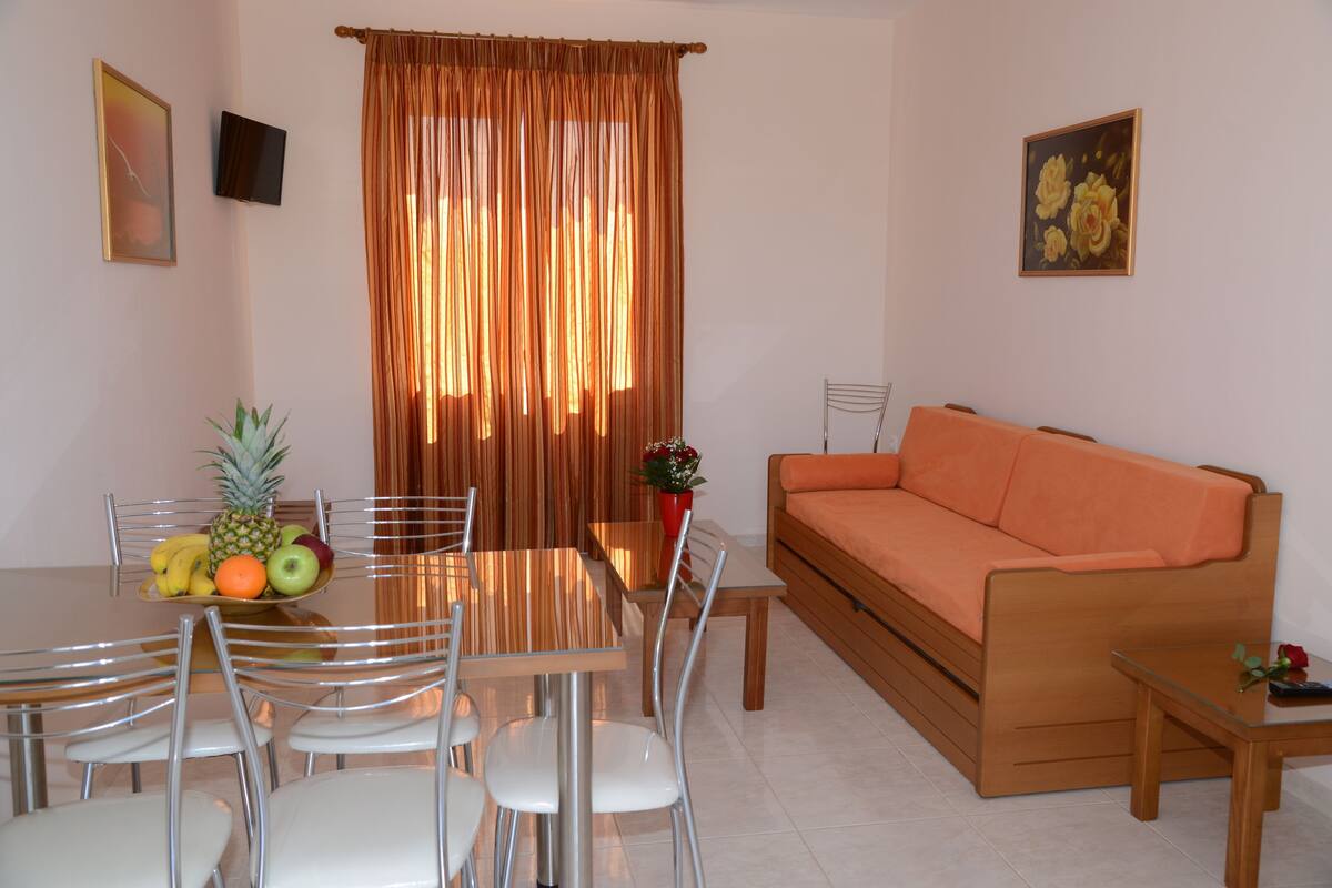 A dining area with a table, chairs, and a fruit bowl. There is a sofa and a small table with a rose on it.