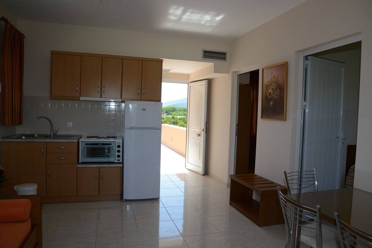 A compact kitchen with wooden cabinets, a built-in oven, and a refrigerator. There is a dining area with a table and chairs, and a view of the outdoors through an open door.