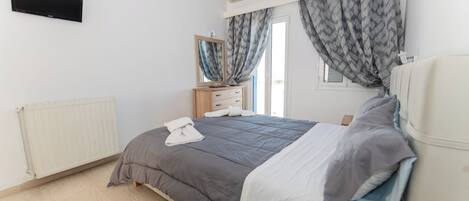 A bedroom with a bed, a dresser, a mirror, a flat-screen TV, and a window with curtains.