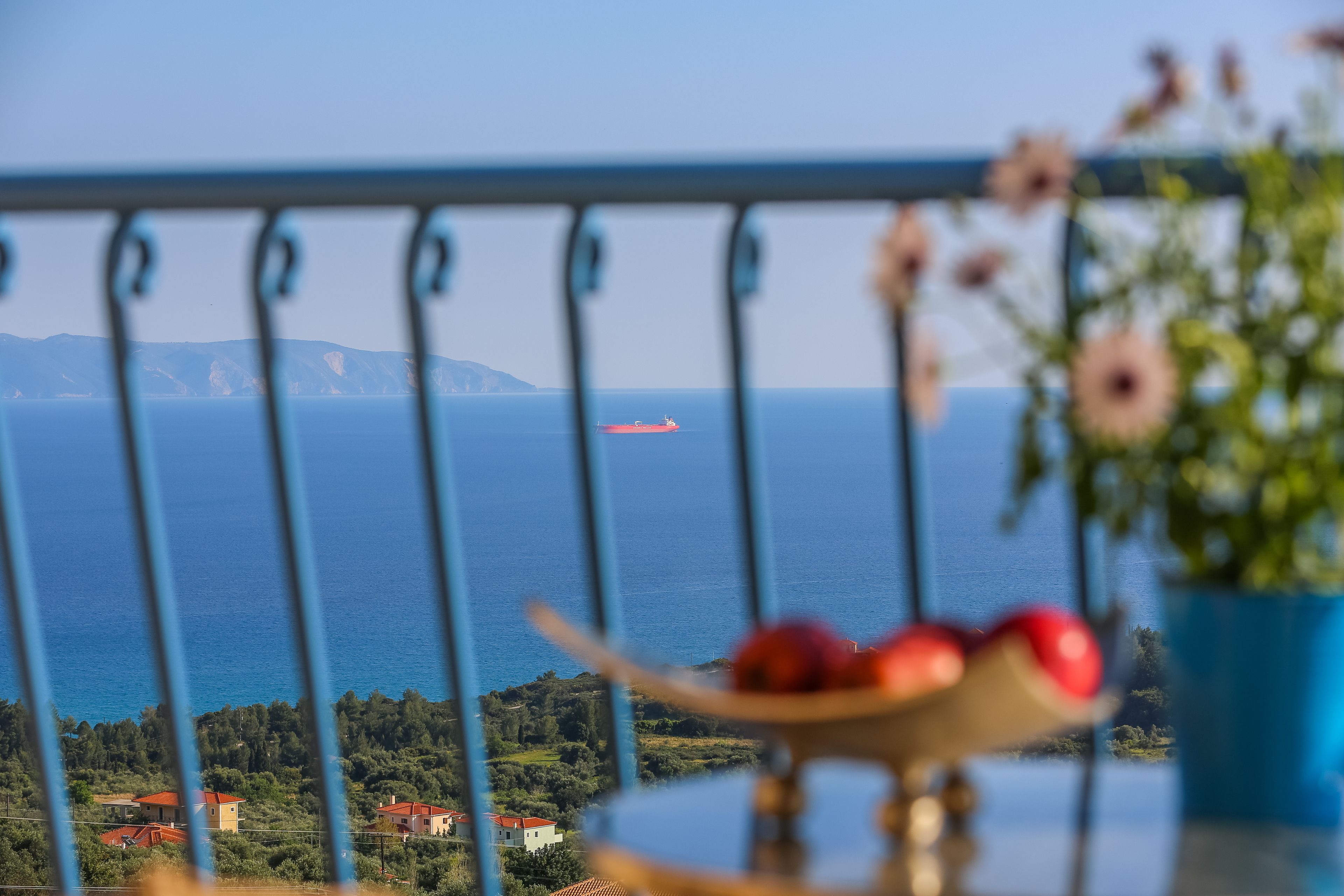 A balcony with a railing, a table with a fruit bowl, and a view of the sea and distant buildings.