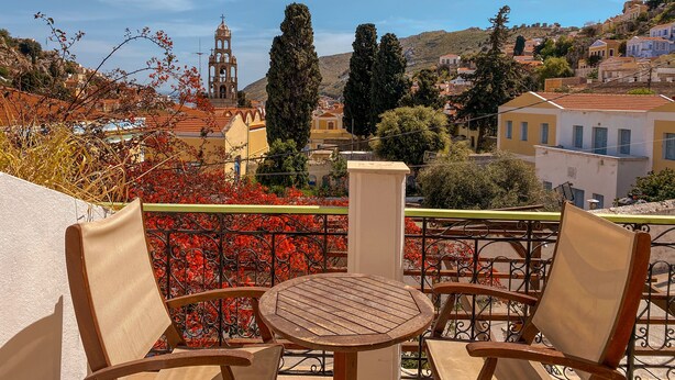 A terrace with two wooden chairs and a table, overlooking a hillside with buildings and a bell tower. - Symi Garden Studios (Symi)