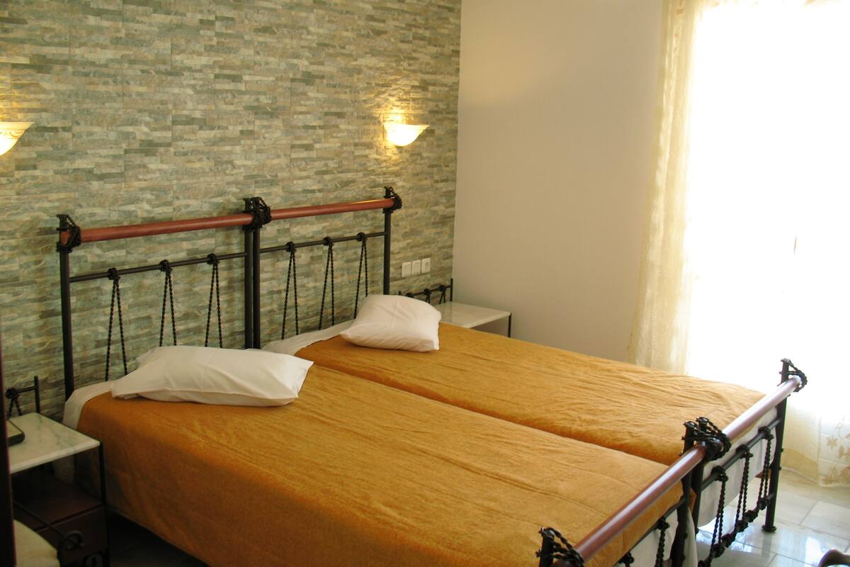 A double bed with a wooden frame and a metal headboard, two pillows, a bedside table, and a wall with a patterned design.