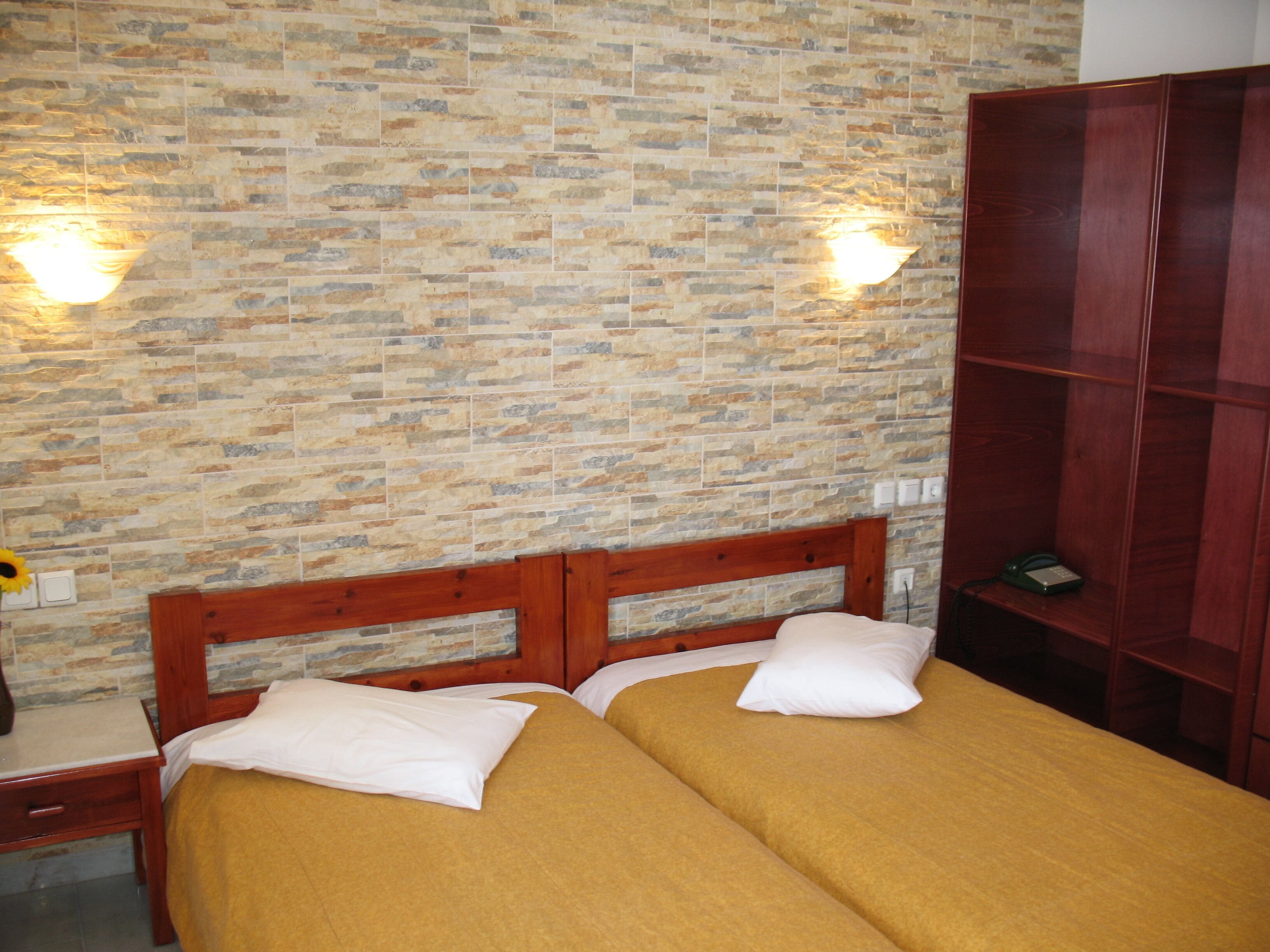 Habitación con dos camas, cabeceros de madera, pared con estampado de ladrillo y un armario de madera.