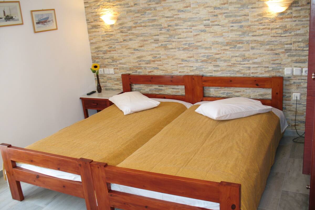A double bed with wooden headboard and footboard, two white pillows, and a wooden nightstand with a vase of sunflowers.