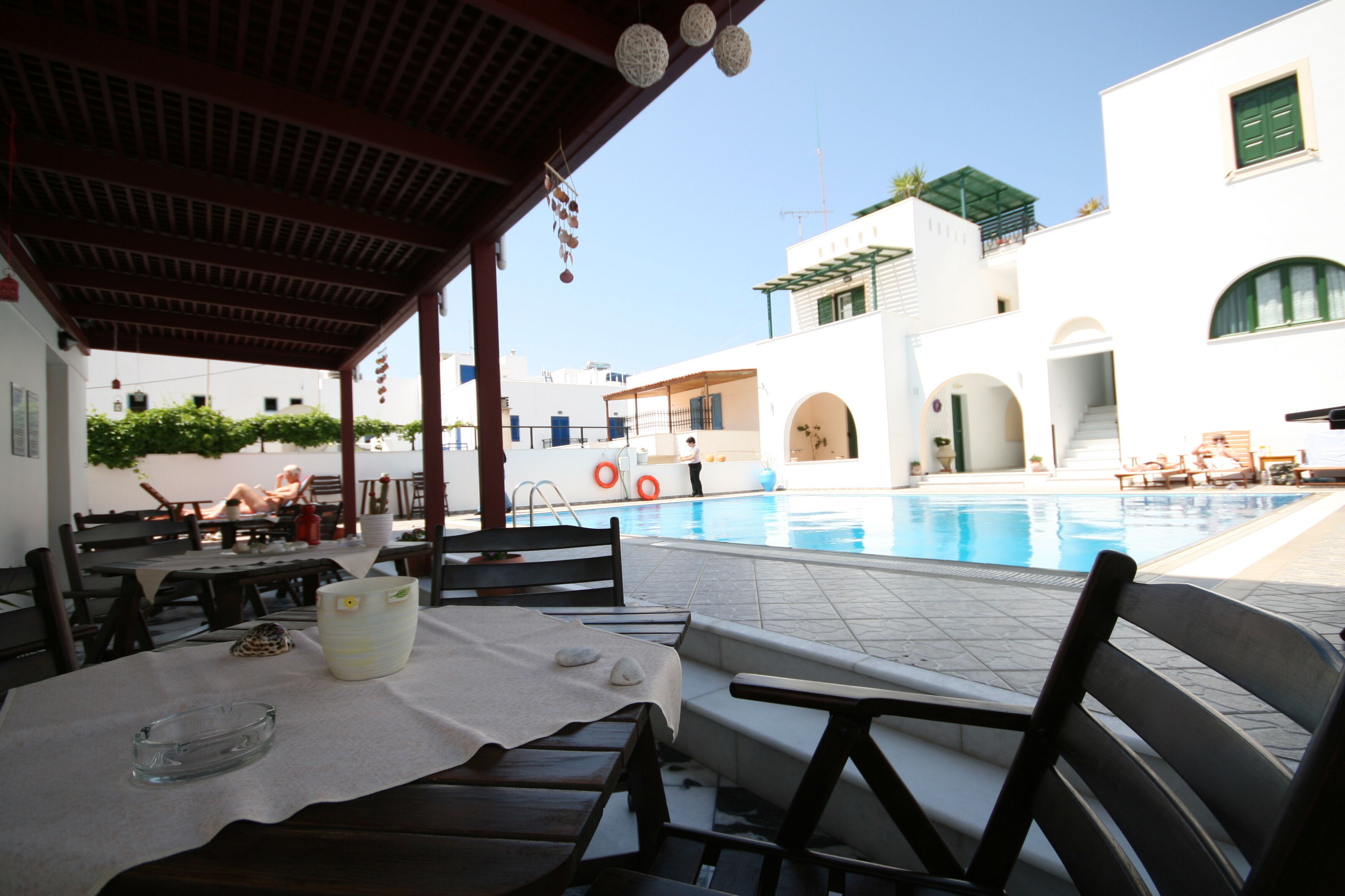 A poolside area with a covered seating area, a pool, and a building with white walls and green shutters.