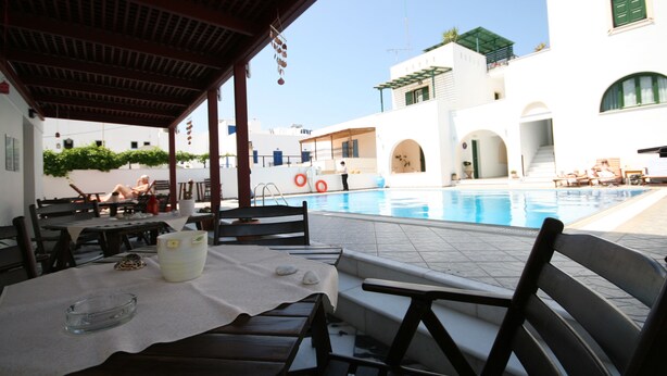 Un área junto a la piscina con una zona de estar techada, una piscina y un edificio de paredes blancas y contraventanas verdes.