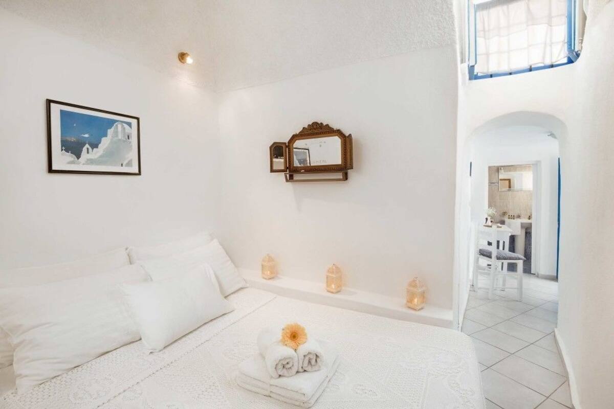A white bedroom with a bed, pillows, a framed picture, a mirror, and a small table with a towel and flower.