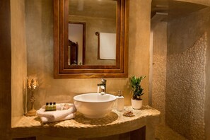 A bathroom with a large mirror, a sink, a towel, a plant, and a stone-textured wall. - Annio Flats (Santorini)