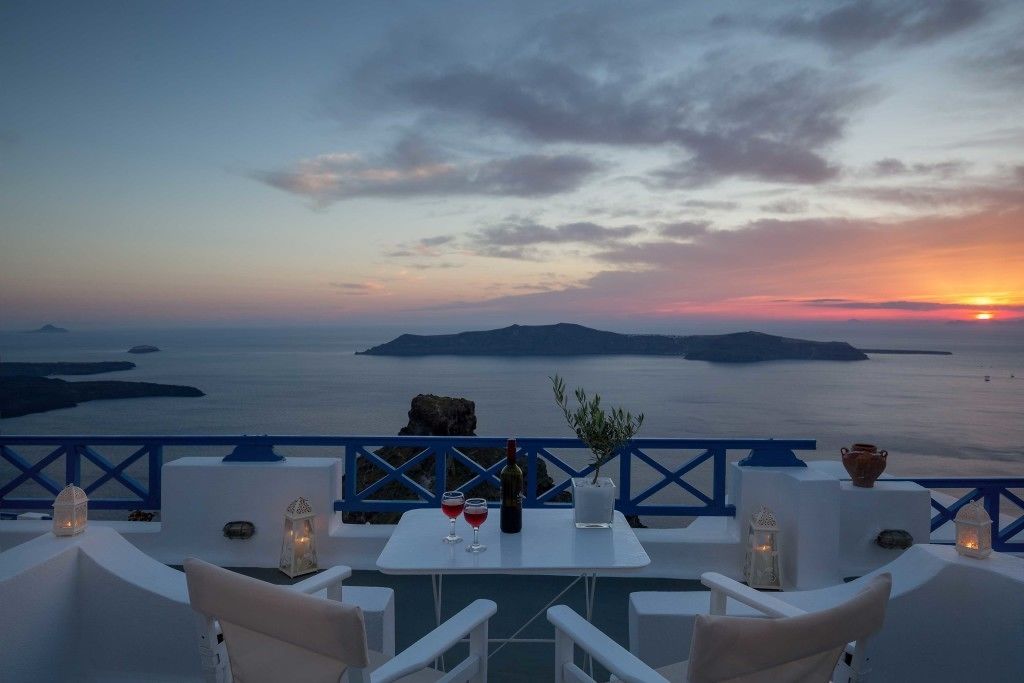 Une terrasse avec des meubles blancs, une table avec une bouteille et des verres, et une vue sur la mer et les îles au coucher du soleil.