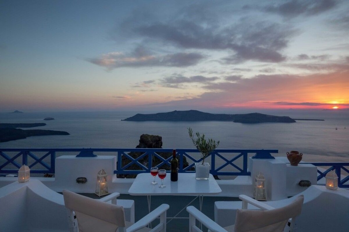 A terrace with white furniture, a table with a bottle and glasses, and a view of the sea and islands at sunset.