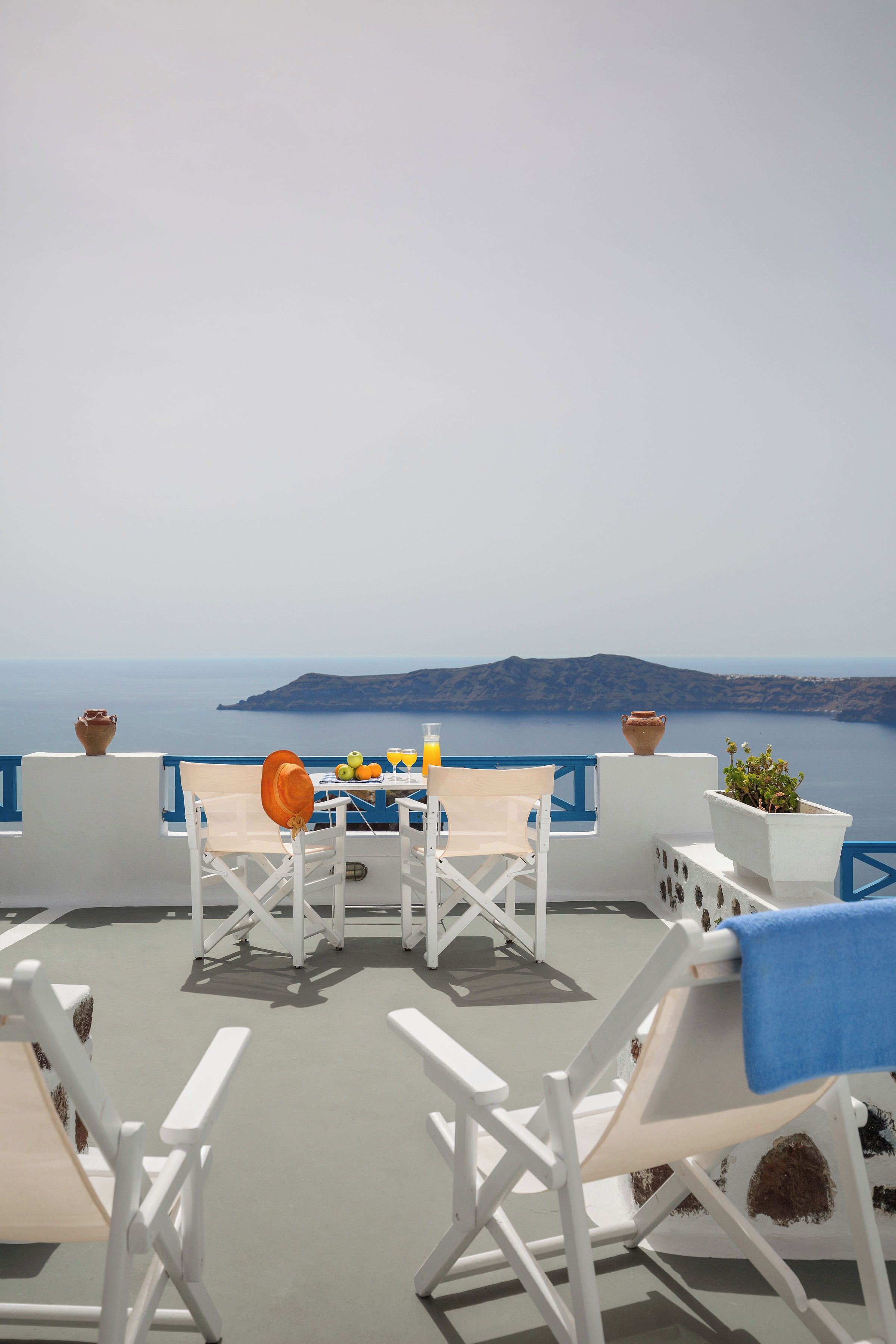 Une terrasse avec des meubles blancs, une table garnie de fruits et de boissons, et une vue sur la mer et les collines.