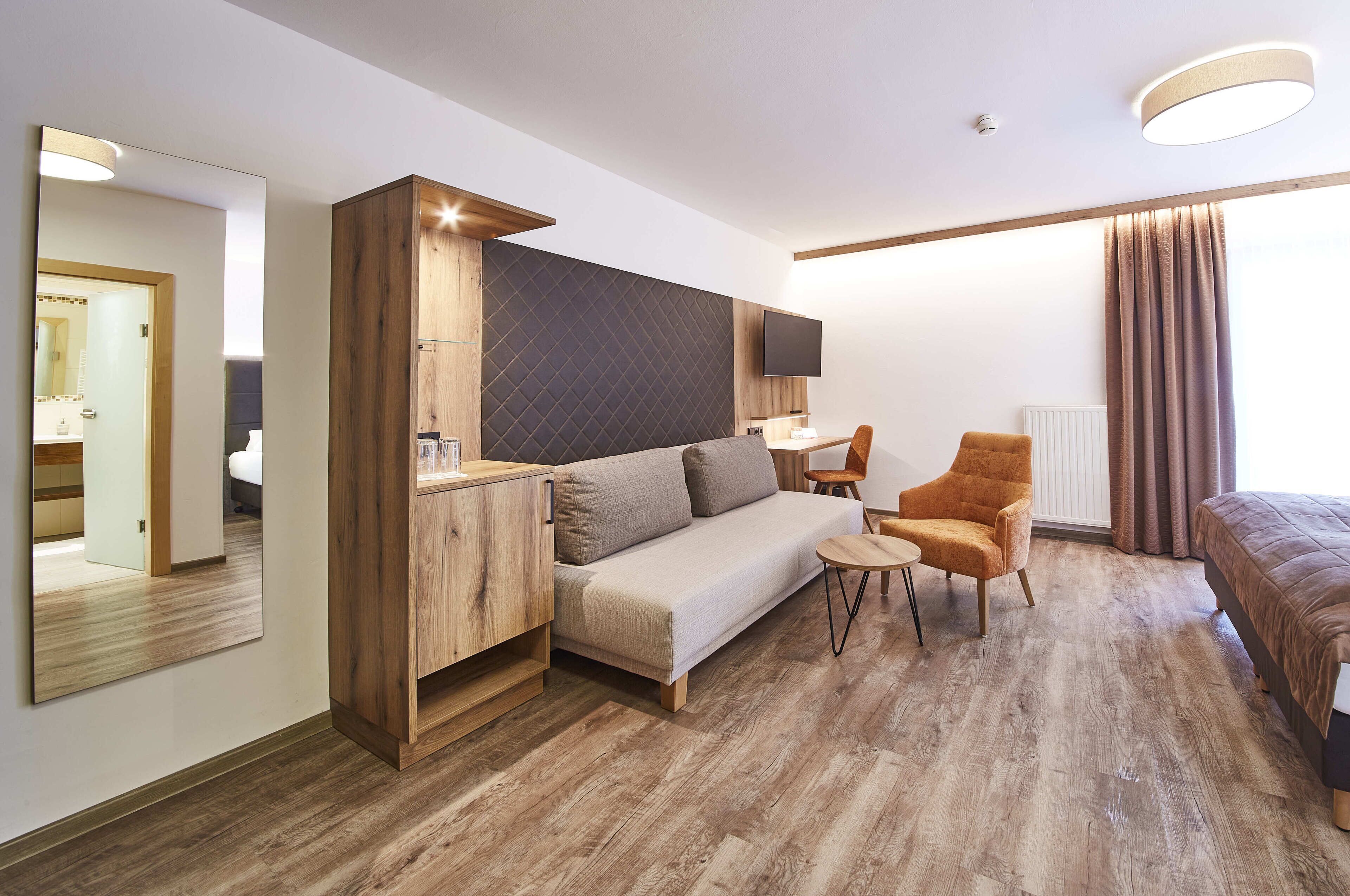 A modern hotel room with a wooden floor, a sofa, a small table, a chair, a bed, a TV, and a large mirror.