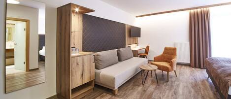 A modern hotel room with a wooden floor, a sofa, a small table, a chair, a bed, a TV, and a large mirror.