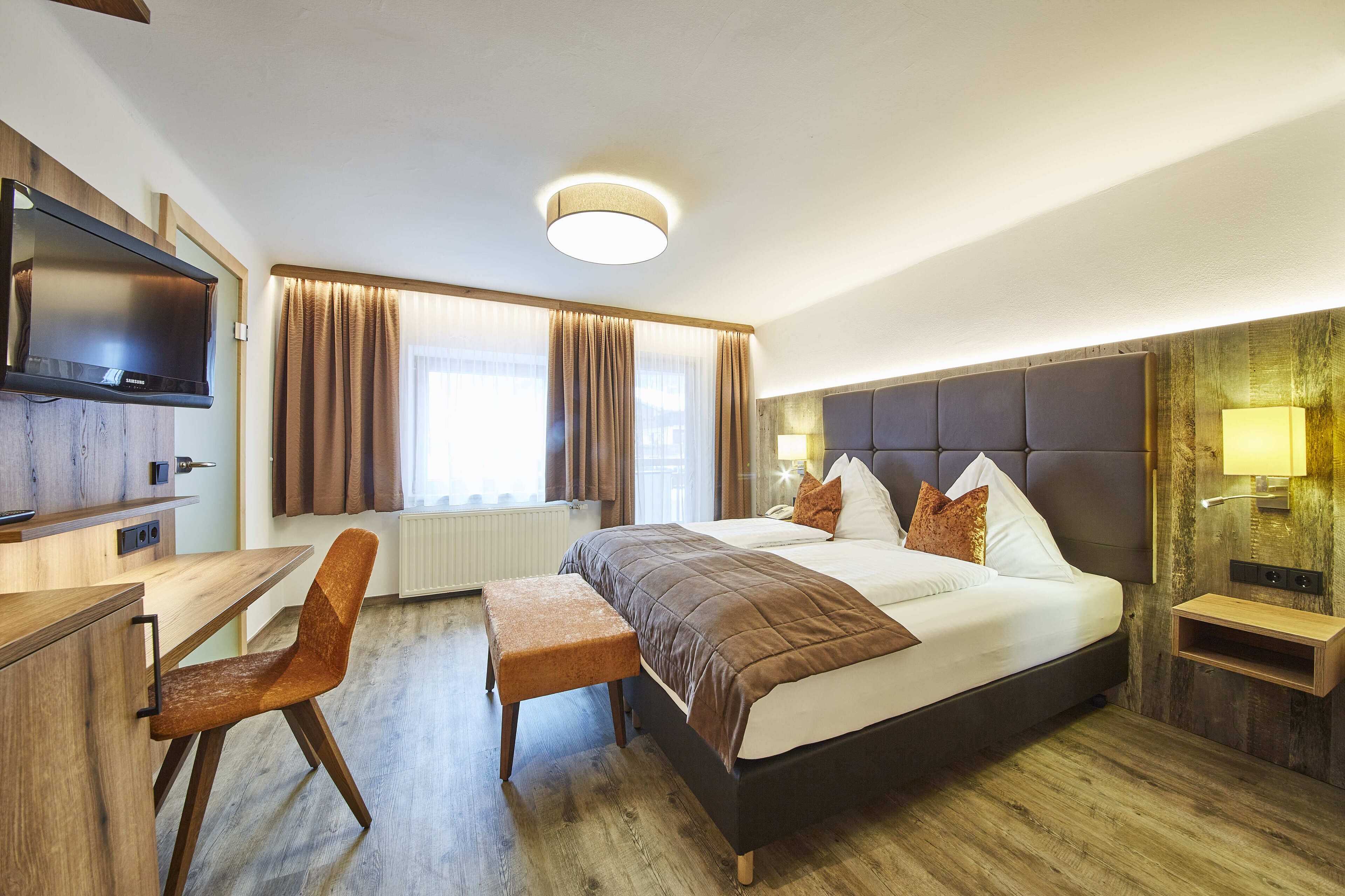 Una habitación de hotel moderna con una cama grande, un escritorio con silla, televisor y ventana con cortinas.