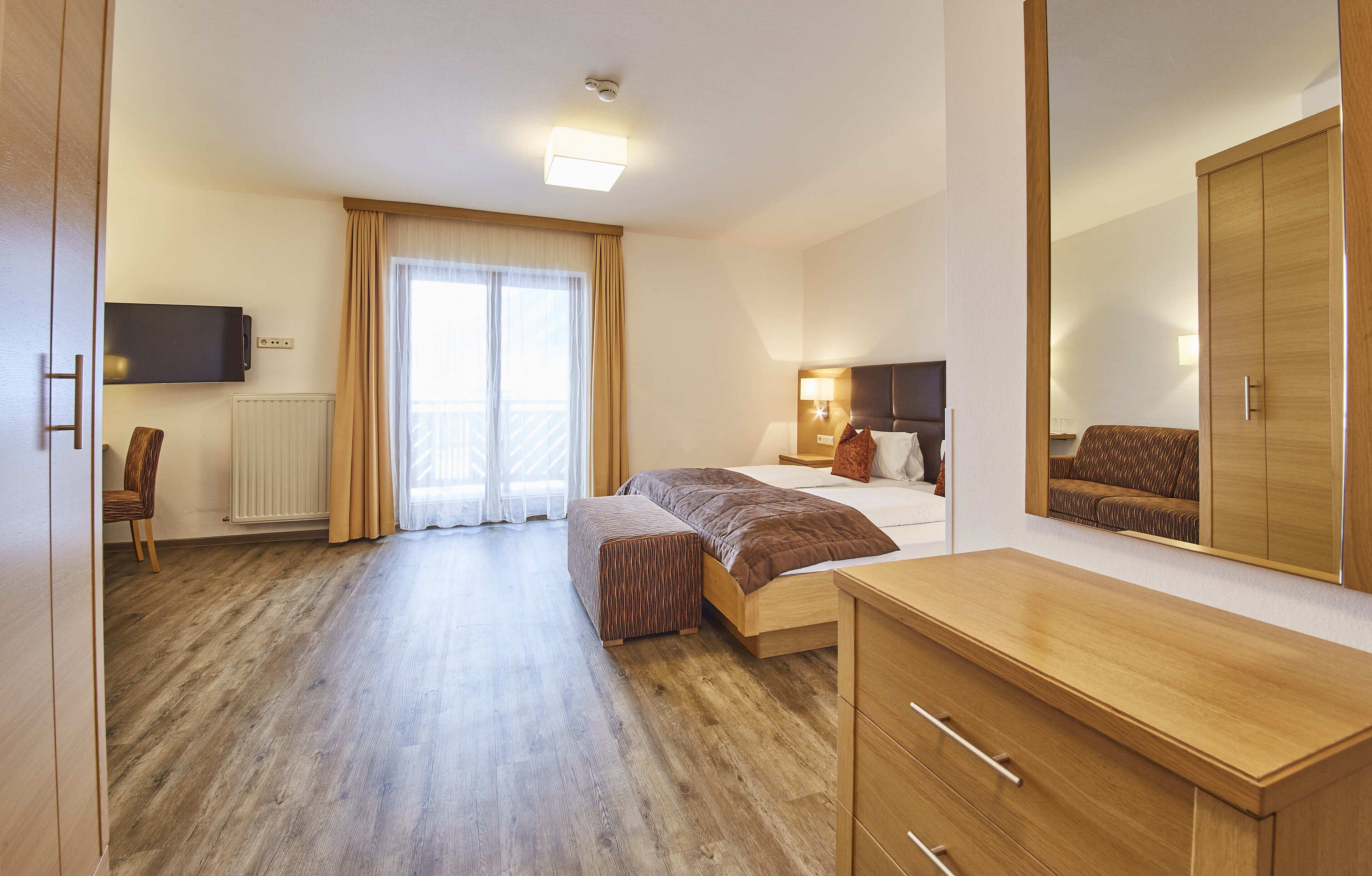 A hotel room with a wooden floor, a bed with a brown headboard, a bedside table, a mirror, and a television mounted on the wall.