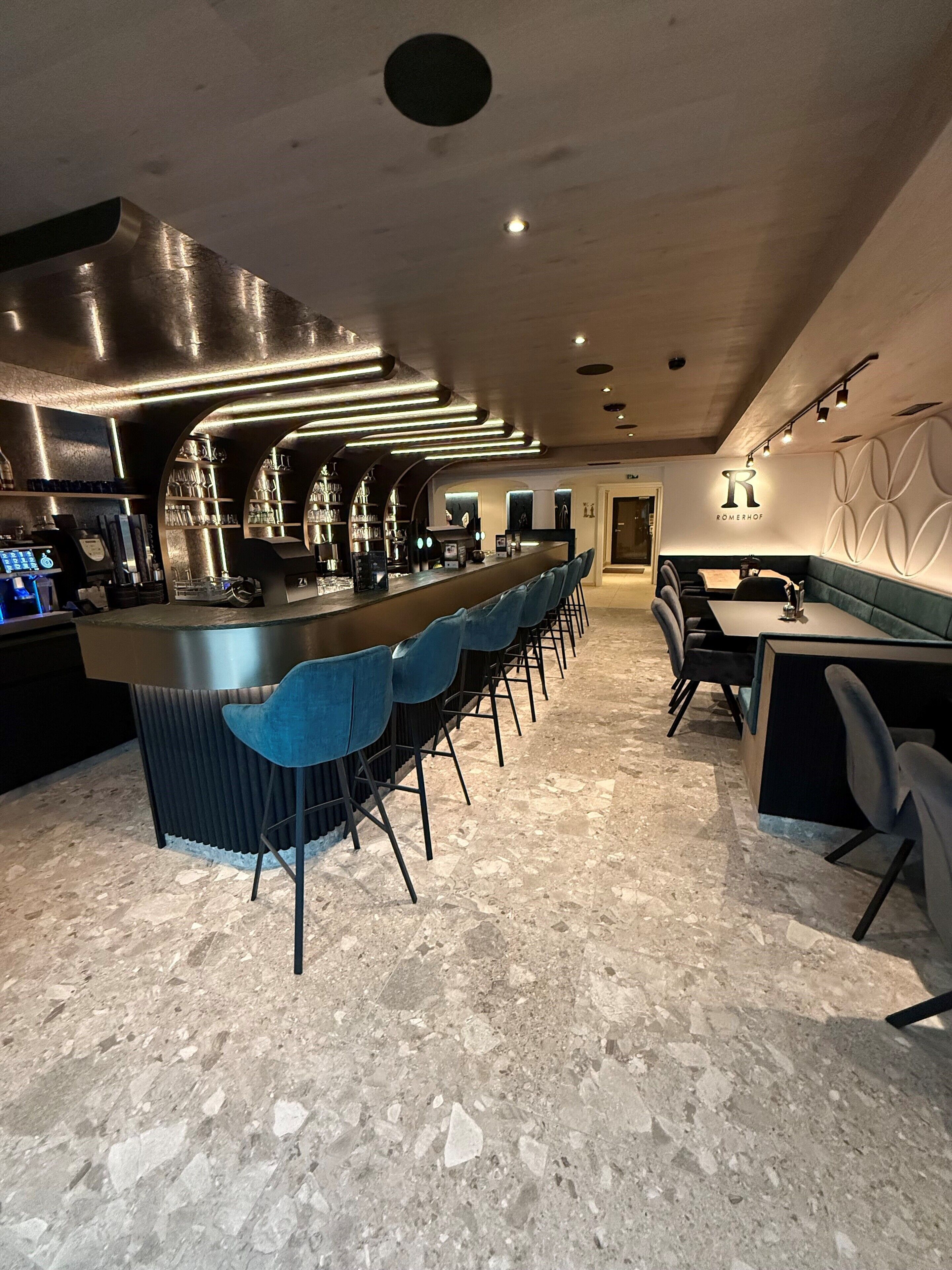 A modern bar area with a long counter, blue bar stools, and a seating area with a table and chairs.