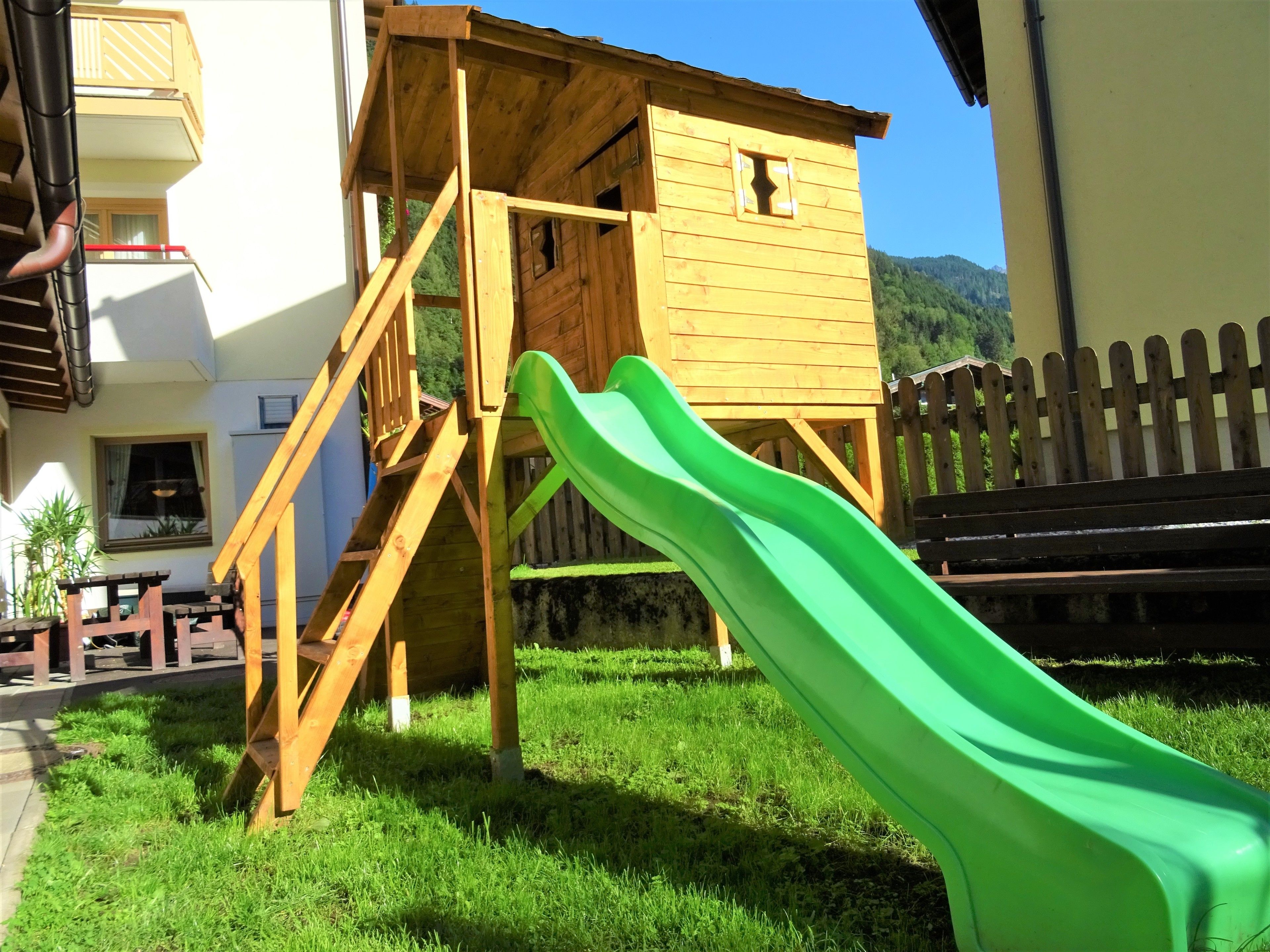 A wooden playhouse with a slide, a staircase, and a fenced area.