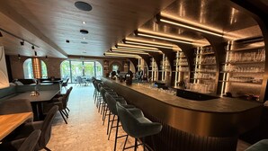 A modern bar area with a long counter, bar stools, and shelves of glasses. - Hotel Römerhof (Fusch an der Grossglocknerstrasse)