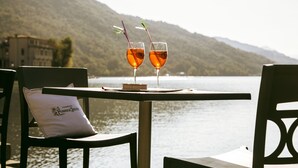Dos vasos de líquido ámbar sobre una mesa con un cojín que dice "Champagne Perrier-Jouët" y una vista de un lago con montañas al fondo.