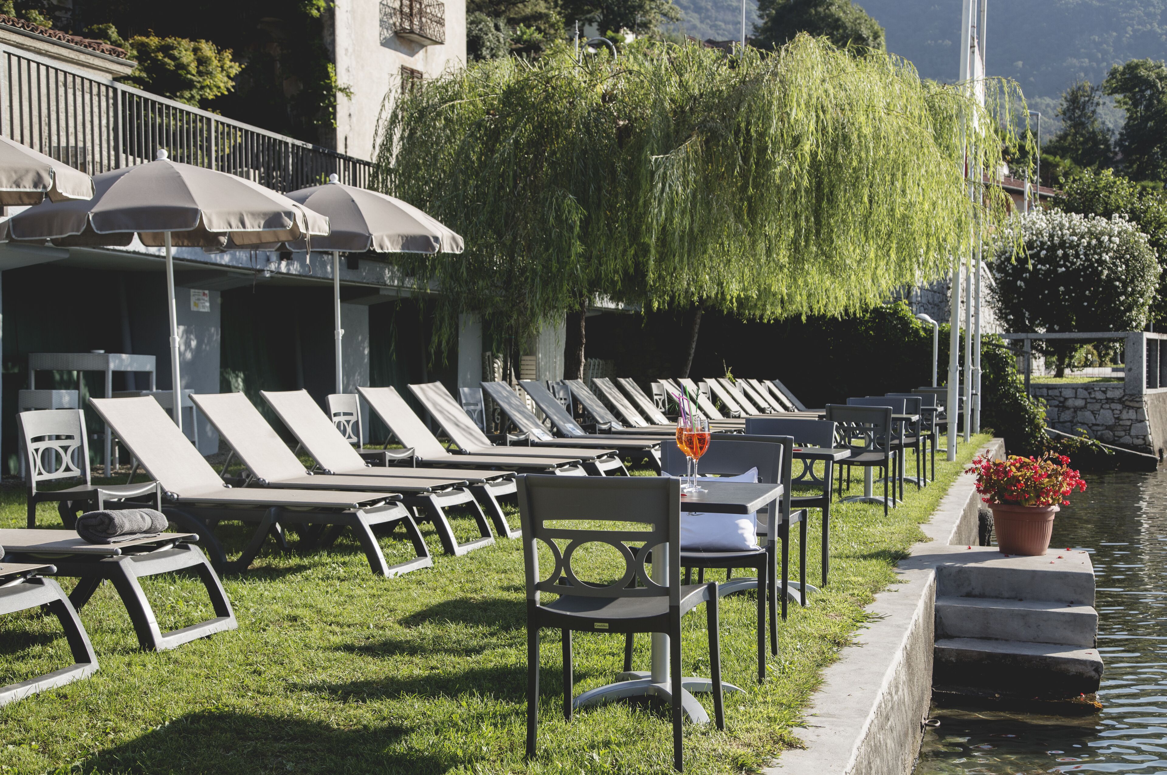 Ein Poolbereich mit Liegestühlen und Sonnenschirmen, ein kleiner Tisch mit einem Getränk und ein Blumentopf am Wasser.