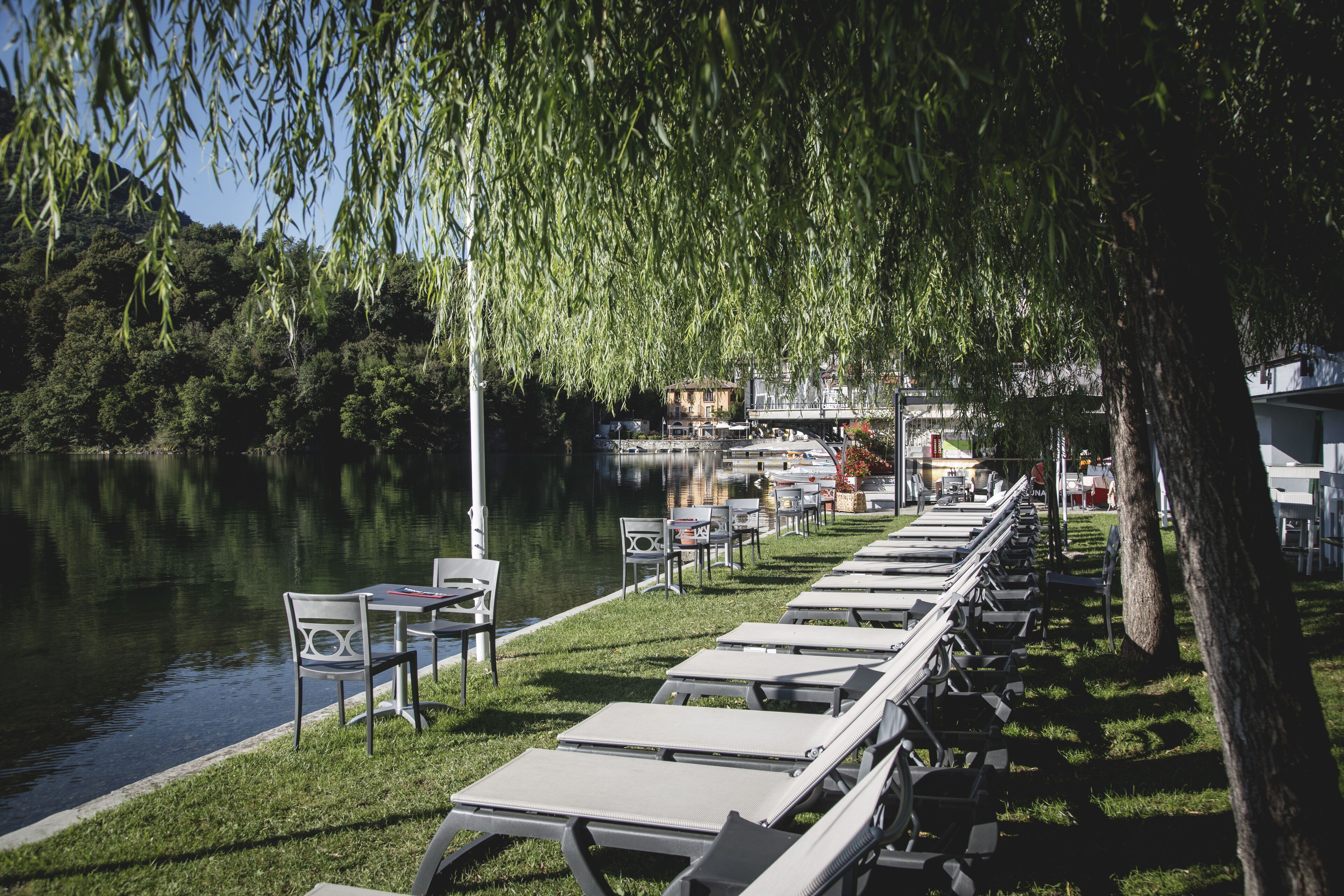Un espace aménagé au bord du lac, avec des tables et des chaises, une pelouse verdoyante et une vue sur un bâtiment situé de l’autre côté de l’eau.