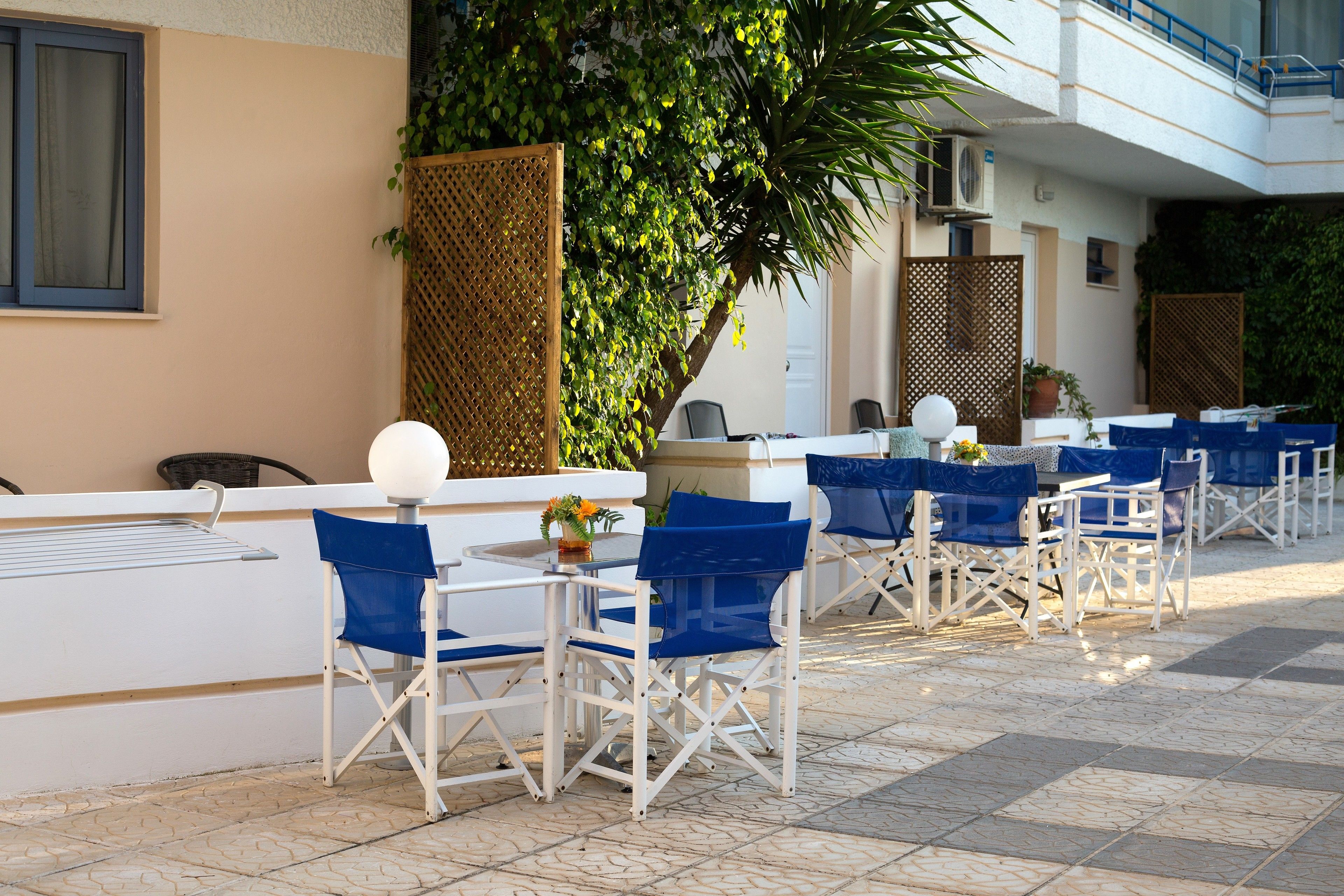 An outdoor seating area with white tables and blue chairs, a large tree, and a building with a balcony.