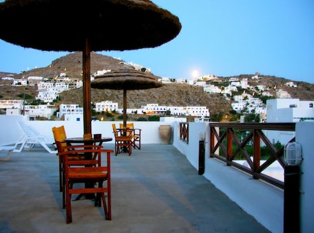 A terrace with a table and chairs, a thatched umbrella, and a view of white buildings on a hillside.