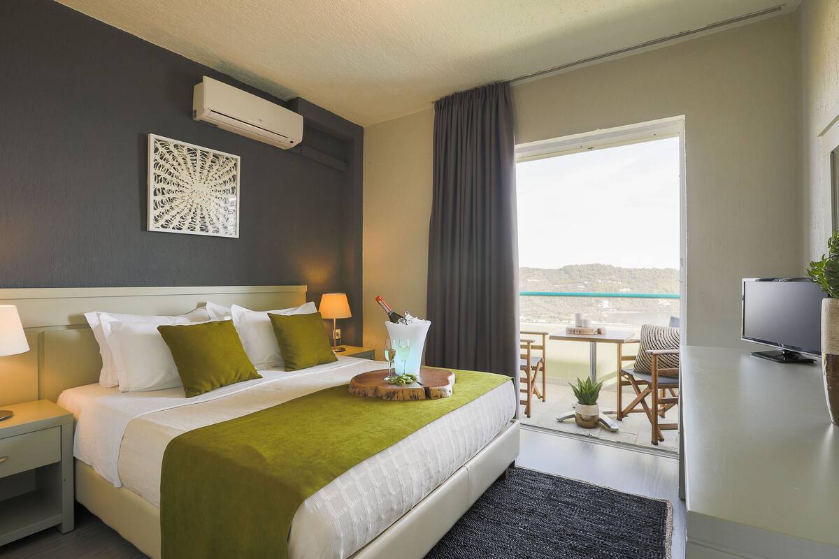 Une chambre d’hôtel avec un lit, une télévision, un balcon avec une table et des chaises, et une vue sur la mer.