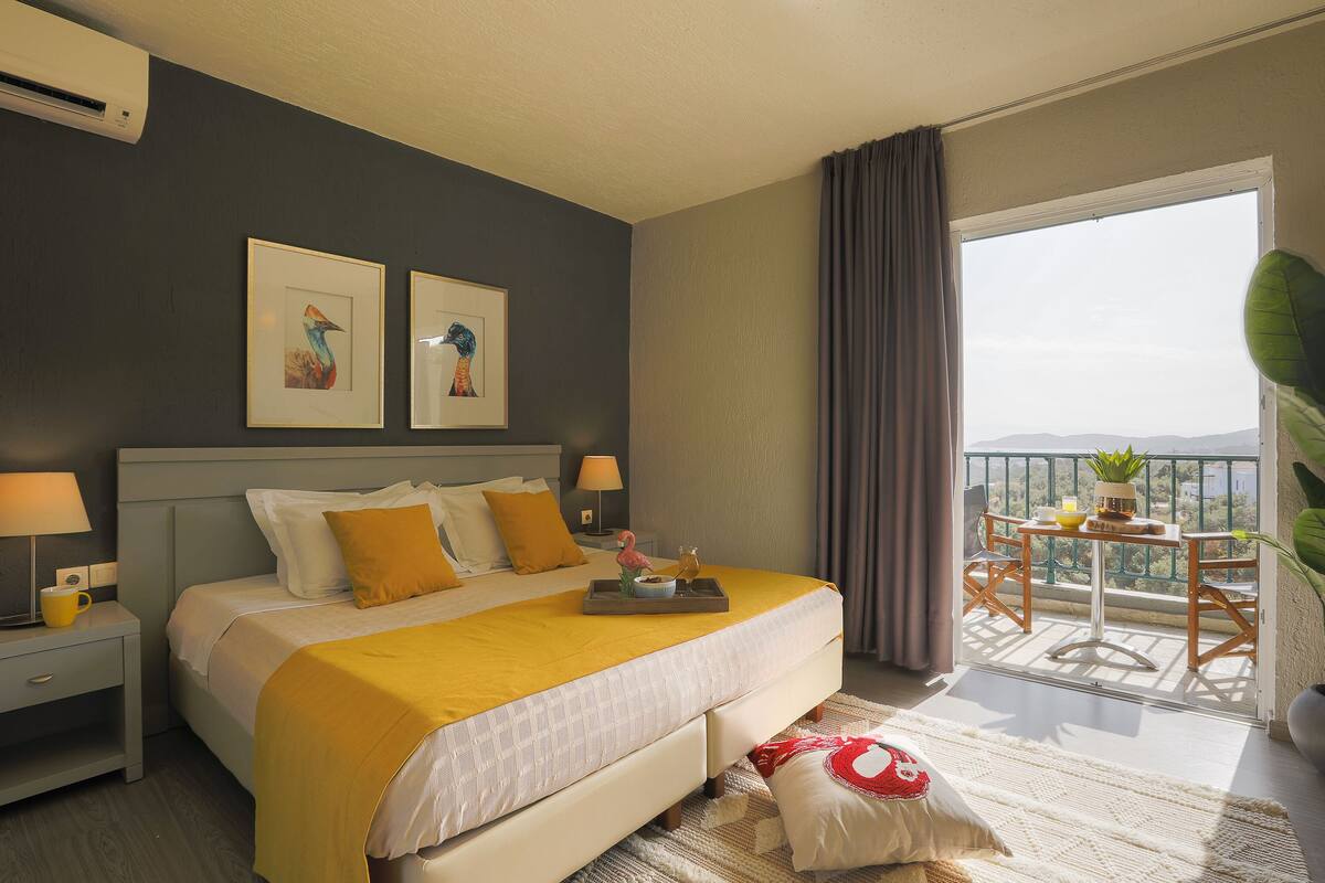 Une chambre d’hôtel comprenant un lit, une table de chevet, une commode, une fenêtre avec des rideaux et un balcon avec une table et des chaises.