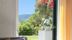A room with a sofa, a small table with books, and a view of a garden with red flowers and a mountain in the background. - Vigles Sea View (Skiathos)