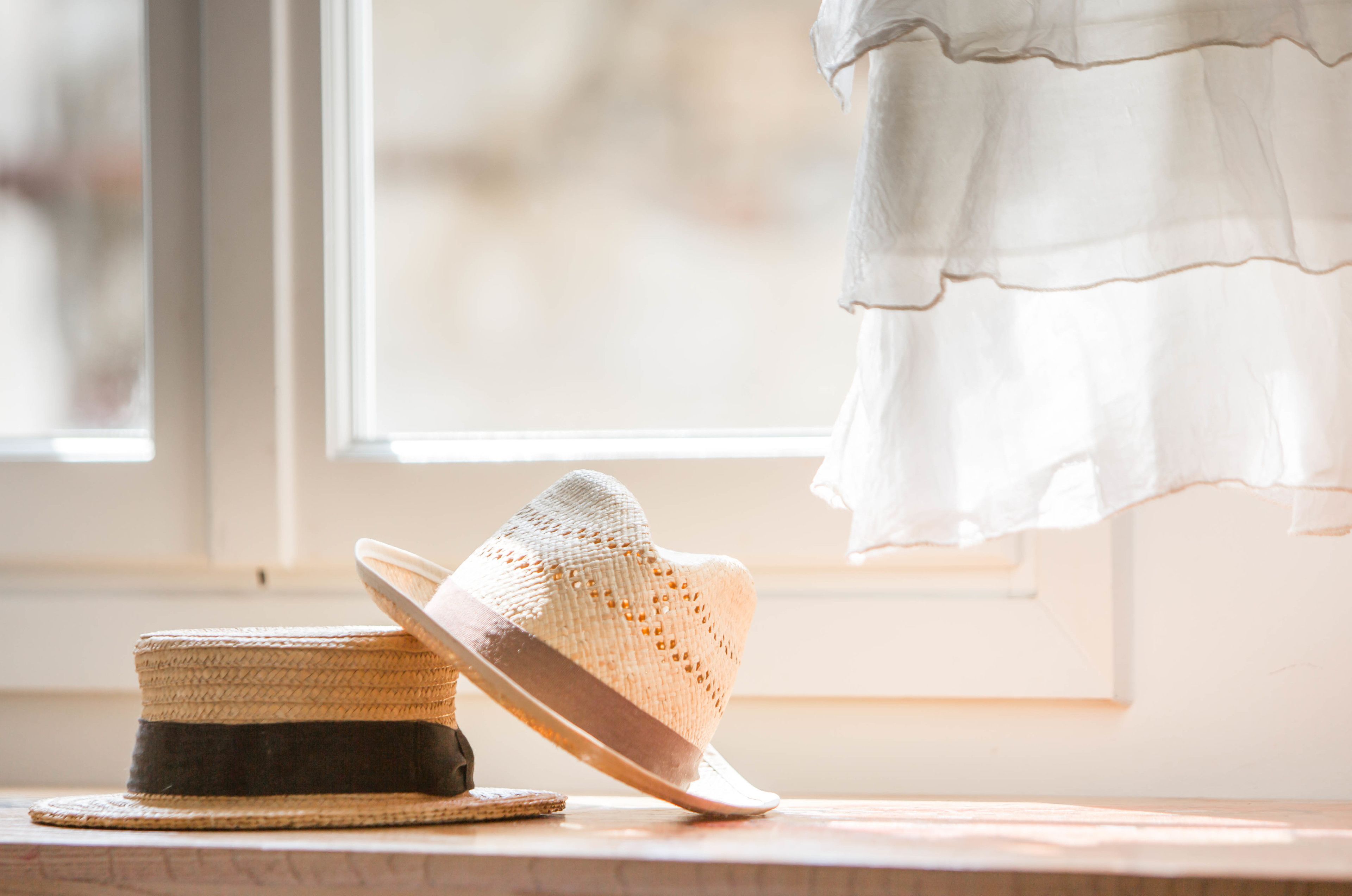 A straw hat and a woven basket on a wooden surface near a window with a white curtain.