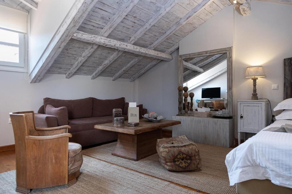 A cozy room with a sloped ceiling, a brown sofa, a wooden coffee table, a small ottoman, and a bed with white bedding.