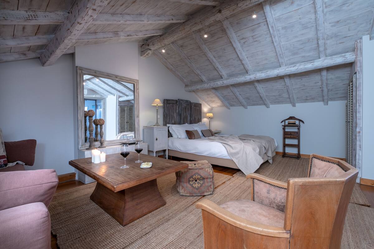 A cozy bedroom with a wooden ceiling, a bed with a white duvet, a nightstand with a lamp, and a window with a view.
