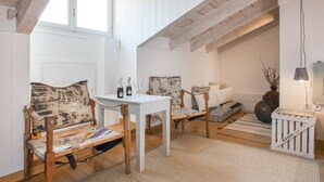 Una habitación acogedora con techo de madera, paredes blancas y una pequeña mesa con botellas y vasos. Hay dos sillas de madera y un banco con cojines. Un jarrón y una lámpara están sobre el suelo.