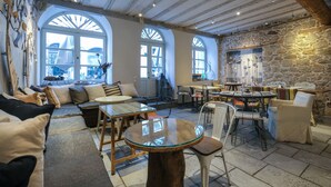 Una habitación amplia con techo alto, paredes de piedra y ventanas arqueadas grandes. Cuenta con un comedor con una mesa redonda y sillas, y un espacio de descanso con sofás y cojines.